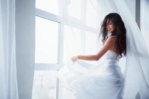 dietro le tende. bella donna in abito bianco si trova in una stanza bianca con la luce del giorno attraverso le finestre foto