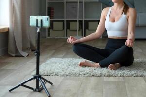 donna fare yoga a casa su il tappeto, seduta nel loto posizione con sua Telefono e tripode per registrazione S o vivere streaming per un in linea fitness centro classe attraverso un' sociale media piattaforma. foto