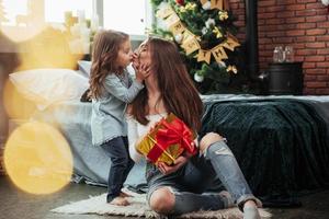 simpatica scena di bacio. madre e figlia siedono in una stanza decorata per le vacanze e tengono in mano una confezione regalo