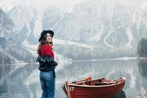 vista spettacolare. sorridendo per la foto. donna con cappello nero che si gode il maestoso paesaggio di montagna vicino al lago con la barca foto