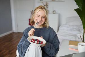 ritratto di carino sorridente biondo donna mangia prima colazione nel sua Camera da letto, guardare a telecamera, Tenere ciotola con dolce e cucchiaio foto