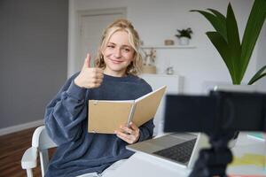 sorridente, positivo giovane donna, Spettacoli pollici su, si siede nel camera con digitale telecamera e computer portatile, record , dà in linea esercitazione, crea soddisfare per seguaci foto