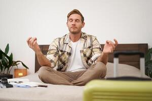 vicino su ritratto di Meditare turista, uomo si siede con valigia nel Camera da letto, pratica yoga, rilassante, scoperta nirvana foto