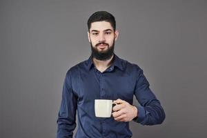 bell'uomo barbuto con capelli alla moda barba e baffi sul viso serio in camicia che tiene tazza bianca o tazza bevendo tè o caffè in studio su sfondo grigio