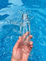 mano spruzzi acqua a partire dal chiaro bicchiere bottiglia in un' blu piscina, dinamico movimento congelare sparo. pulito acqua concetto. foto