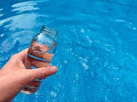 mano Tenere chiaro bicchiere bottiglia parzialmente pieno con acqua contro scintillante blu acqua sfondo con vuoto spazio per testo. pulito acqua concetto. foto