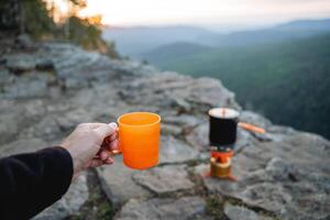 un' mano detiene un' boccale di tè contro il fondale di il tramonto nel il montagne, un' escursionista potabile un' caldo bere, un sera cena solo, montagna escursioni a piedi avventura nel natura. foto