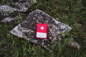 un' rosso primo aiuto kit bugie su un' pietra nel natura, un' piccolo escursioni a piedi Borsa con essenziale medicinali, un' bianca attraversare simbolo, un' medico Borsa. foto