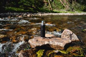 il concetto di turista piatti thermos per tè sta su un' pietra nel il fiume, un' montagna ruscello con freddo acqua, escursioni a piedi nel il montagne, un' fornitura di acqua su il strada foto