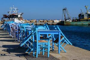 blu di legno tavoli e sedie di un' tipico pesce taverna a il porta. pesca navi su il sfondo. Egeo isola chios nel Grecia su un autunno giorno. greco vacanze e destinazioni foto