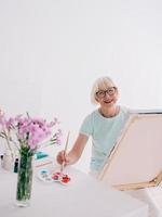 artista senior donna allegra in bicchieri con capelli grigi pittura fiori in vaso. creatività, arte, hobby, concetto di occupazione foto