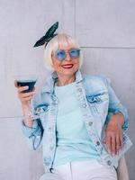 donna anziana alla moda con i capelli grigi e con occhiali blu e giacca di jeans che tiene in mano un bicchiere con cocktail blu. alcol, relax, vacanze, concetto di pensione foto