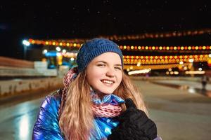 felice sorridente giovane donna caucasica in sciarpa, cappello, giacca, guanti sulla pista di pattinaggio all'aperto. anno nuovo, divertimento, concetto invernale