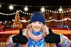 felice sorridente giovane donna caucasica in sciarpa, cappello, giacca, guanti sulla pista di pattinaggio all'aperto. anno nuovo, divertimento, concetto invernale