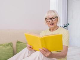 donna anziana con i capelli grigi che legge un libro su un divano a casa. educazione, pensione, anti età, concetto di lettura foto