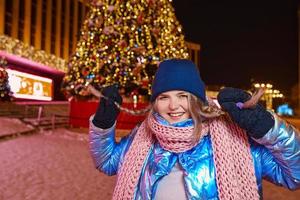 felice sorridente giovane donna caucasica in sciarpa, cappello, giacca, guanti vicino all'albero di Natale all'aperto. anno nuovo, divertimento, concetto invernale