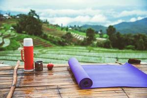 tappetino da yoga viola e bottiglia d'acqua rossa con un bicchiere d'acqua inossidabile proposto. e mela rossa su fondo di legno. sul balcone paesaggio naturale field.travel relax. papongpieng in thailandia foto