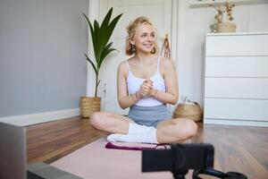 ritratto di giovane fitness istruttore, vlogger mostrando esercizi su telecamera, registrazione se stessa, seduta su stuoia con computer portatile, fare allenamento, spiegando yoga movimenti per seguaci foto