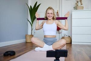 ritratto di giovane sorridente donna, fitness istruttore registrazione di allenamento, mostrando Come per esercizio a casa e uso gomma da cancellare resistenza gruppo musicale, seduta su yoga stuoia foto