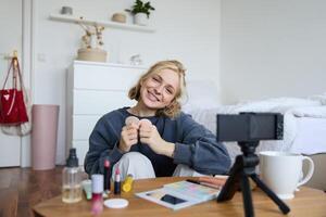 ritratto di sorridente giovane donna, seduta nel davanti di digitale telecamera nel accogliente impostato su, registrazione per stile di vita video blog, chat di trucco e bellezza prodotti foto