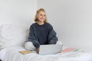 Immagine di contento giovane donna, alunno e-learning a partire dal casa, Collegare per in linea corso su sua computer portatile, si siede su letto con i Quaderni, sorridente e guardare contento foto