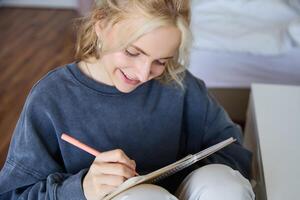 vicino su ritratto di sorridente ragazza, scrittura nel taccuino, Aggiunge Appunti per sua diario, sorridente mentre fare compiti a casa foto