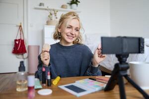 ritratto di giovane donna, bellezza soddisfare Creatore, seduta nel un' camera nel davanti di digitale telecamera, registrazione trucco lezione video blog, mostrando cosmetico facciale prodotti foto