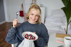 ritratto di sorridente biondo donna, mangiare colazione, Tenere ciotola e cucchiaio, seduta nel Camera da letto, guardare contento a telecamera foto