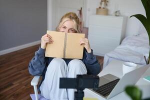 ritratto di carino, sorridente giovane sociale media soddisfare Creatore, ragazza record su digitale telecamera e stabilizzatore, detiene taccuino, parla per pubblico, Vlogging nel sua camera foto