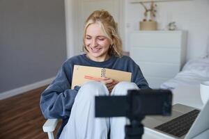 ritratto di sorridente carino donna, stile di vita blogger, si siede nel sua camera con quotidiano rivista o pianificatore, record su digitale telecamera, crea soddisfare per sociale media di quotidiano routine foto