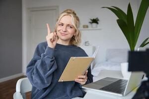 ritratto di giovane creativo donna, soddisfare creatore, seduta nel camera, Lavorando a partire dal casa, utilizzando computer portatile, Tenere taccuino, raccolta dito, ha un idea, eureka gesto foto