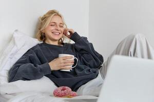 ritratto di contento donna godendo giorno libero a casa, dire bugie nel letto, Guardando tv mostrare su computer portatile, guardare a schermo e sorridente, potabile tè e mangiare nel Camera da letto foto
