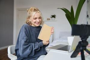 ritratto di carino giovane donna, adolescenziale ragazza record su digitale telecamera, usi il computer portatile per creare stile di vita contenuto, Spettacoli taccuino, legge Appunti, utilizzando computer nel sua camera foto