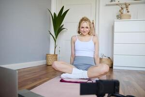 ritratto di giovane fitness istruttore, vlogger mostrando esercizi su telecamera, registrazione se stessa, seduta su stuoia con computer portatile, fare allenamento, spiegando yoga movimenti per seguaci foto