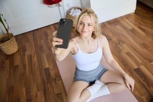 ritratto di elegante, giovane bellissimo donna prende autoscatto mentre Lavorando su a casa, utilizzando smartphone per rendere fotografie su yoga stuoia, in posa nel fitness Abiti