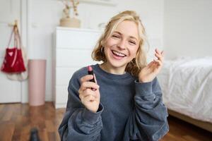 ritratto di giovane sociale media influencer, donna registrazione un' con bellezza prodotti, mostrando sua trucco su telecamera, Tenere rossetto e sorridente, seduta su pavimento foto