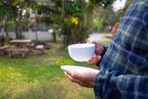 avvicinamento di mani anziano dell'uomo Tenere un' bianca tazza di caffè mentre in piedi nel il giardino. spazio per testo. concetto di anziano persone e rilassamento foto