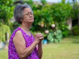 un anziano asiatico donna indossare bicchieri si chiude sua occhi e pregare mentre in piedi nel il giardino. spazio per testo. concetto di anziano le persone, inverno stagione e religione foto