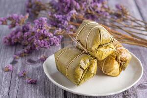 al vapore appiccicoso riso con Banana avvolto Banana foglia su piatto con viola fiori sfocato sfondo. foto