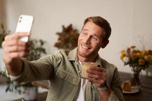 ritratto di sorridente bello giovane uomo, chat con amico a partire dal bar, fare selfie con tazza di caffè foto