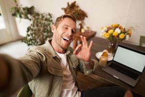 bello sorridente giovane uomo assunzione autoscatto, mostrando va bene bene mano cartello, Fotografare su mobile Telefono, si siede nel bar con il computer portatile foto