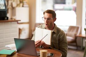 ritratto di bello giovane uomo dando Lezioni in linea, mostrando esercizi nel il suo taccuino, guardare a computer portatile, registrazione , seduta nel bar, abbastanza co-working spazio foto