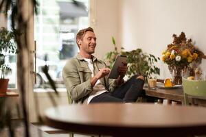 ritratto di bello contento tipo, giovane uomo si siede nel bar, Guardando S su digitale tavoletta, indossare senza fili cuffia, ridendo e sorridente, la spesa tempo nel caffè negozio foto