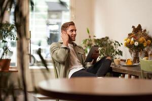 ritratto di bello sorridente giovane uomo, seduta nel caffè negozio, utilizzando digitale tavoletta, chat con qualcuno a partire dal un' bar, indossare senza fili cuffie foto