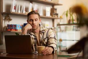 ritratto di uomo guardare triste e stressato a il suo bicchiere di caffè, Lavorando a partire dal bar, seduta con computer portatile, manca ispirazione, no sentire ispirato per finire il progetto foto