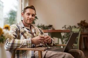 Immagine di bello uomo d'affari Lavorando a partire dal bar, potabile caffè e fare in linea progetto su il suo computer portatile, sorridente e guardare felice, finito il suo libero professionista compito, guardare lieta foto