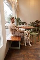 verticale tiro di giovane uomo, proprietario di d'oro cane da riporto, seduta nel bar con e accarezzando il suo cane sotto il tavolo, potabile caffè nel un' caffetteria foto