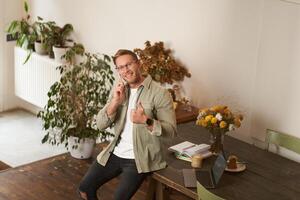 ritratto di bello contento uomo nel un ufficio, tipo si siede nel coworking spazio, guardare a telecamera, sorridente e ridendo, parlando su mobile Telefono, utilizzando smartphone, risposta un' chiamata a partire dal cliente foto