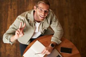 superiore angolo tiro di bello sorridente uomo nel occhiali, mostrando pace cartello e sorridente a telecamera, Lavorando nel bar, studiando nel davanti di computer portatile, lettura Appunti nel taccuino foto