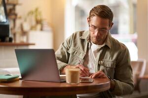 Immagine di focalizzata giovane uomo nel occhiali, seduta nel bar, fabbricazione Appunti, studiando, frequentando in linea corso, apprendimento su a distanza a partire dal abbastanza caffè negozio foto